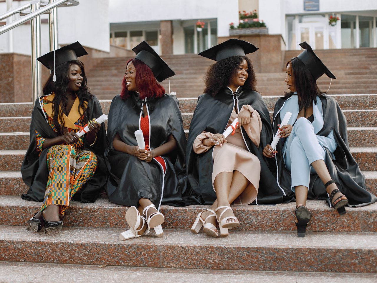 Group of young afro american female students dressed in black graduation gown. Campus as a background. Girls sitting on a stairs and talking.